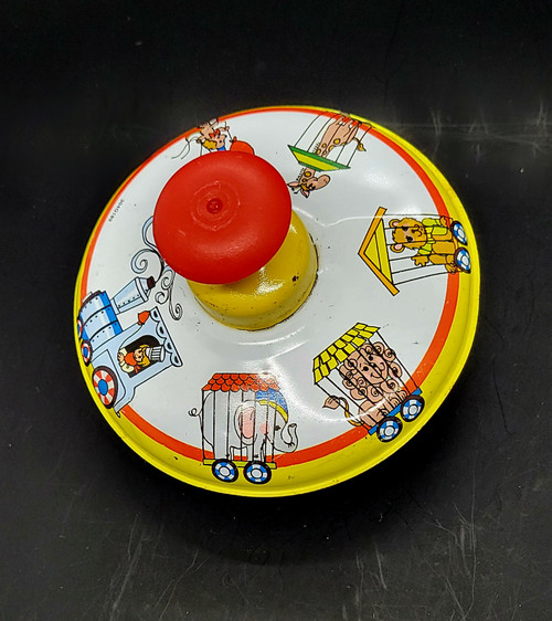 Ohio Art toy spinning top with circus train lithographs that is 5.25-inches diameter. The circus train features conductor pulling 4 cars with elephant, lion, bear and giraffe in the caged train cars. There is a dog with balloons on unicycle following the train.