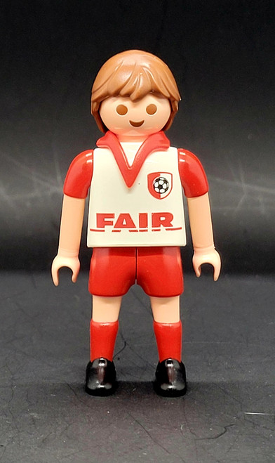 Geobra the German manufacture of Playmobil 2006 white jersey footballer / soccer player from the Playmobil 4700 Soccer Football Stadium set. The figure is wearing a white jersey with red sleeves, shorts and socks. The jersey also has FAIR in red letters across the front and 23 as soccer jersey number. The player has lever to create a kicking motion.