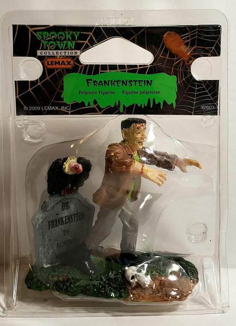 Lemax Spooky Town Frankenstein Figure #92609, a figurine of Frankenstein with a scarred face, dressed in a brown jacket and gray pants, walking on a patch of ground next to a tombstone and skulls, with a rat crawling on the tombstone.