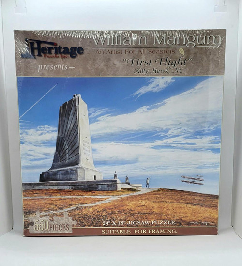 Heritage Puzzles Heritage 550 Piece Jigsaw Puzzle: First Flight Kitty Hawk, NC, featuring an image of the Kitty Hawk monument against a blue sky with clouds and planes flying in formation.