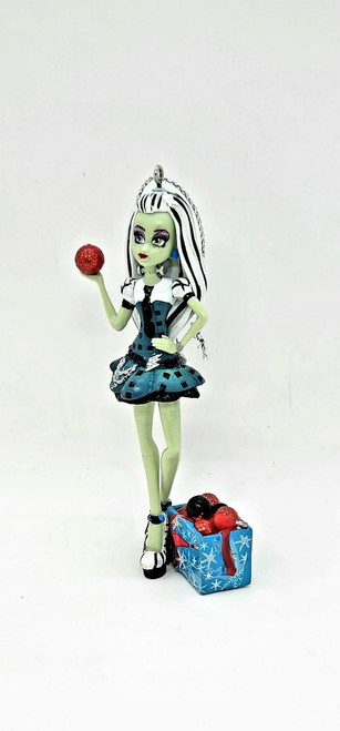 Monster High - Frankie Stein Ornament (2014), detailed green-skinned doll with black and white hair, dressed in a blue and black dress, holding a red ornament, standing next to a blue gift box filled with red and black berries, decorative and collectible Halloween ornament.