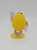 M&M's 2018 Yellow M&M Character Case Plastic Figure, a yellow egg-shaped toy with arms, legs, white gloves, and sneakers, standing on a yellow base and waving.