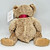AVON 2002 Celebrates The Year of the Teddy Bear Talking Bear, a plush teddy bear with a beige, fluffy fur texture, sitting upright with a red interior in its open mouth, and a tag attached to its left ear.