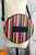 The Oblong Box Shop Haunted Holiday Cat Crossbody Purse, round purse with colorful vertical stripes in black, white, orange, green, and red, featuring a black leather patch with embossed logo.