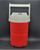 Igloo Laguna 1/2 gallon water cooler jug that is red with white top. The cooler has hooked handles that can hang on a chain link fence. It features a white swing-up carry handle and a secure flip-cap for dispensing.