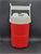 Igloo Laguna 1/2 gallon water cooler jug that is red with white top. The cooler has hooked handles that can hang on a chain link fence. It features a white swing-up carry handle and a secure flip-cap for dispensing.
