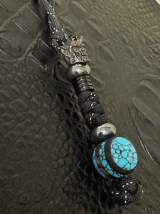 Handmade Turquoise Orbital Bead Knife Lanyard with Black Samurai Mask, Hematite Accents, and Black Metal Column Bead on Reflective Black Paracord