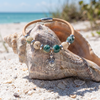 Cork bracelet with green and cream ceramic beads and sterling silver turtle charm displayed in a seashell on the beach — Crabby Mermaid