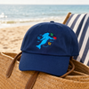 Crabby Mermaid original hat in navy resting on a woven beach bag with ocean in the background - Crabby Mermaid