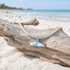 Larimar whale tail cork necklace resting on driftwood with a second driftwood piece and shoreline in the background