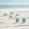 Silver-toned crab, dolphin, and shell stud earrings with light blue enamel displayed on weathered white wood with sand and ocean background — coastal earring set — Crabby Mermaid