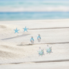 Silver-toned starfish, flip flop, and sea turtle stud earrings with blue enamel and crystal accents displayed on weathered white wood with sand and ocean background — coastal earring set — Crabby Mermaid