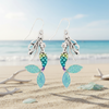 Silver-toned glittering mermaid dangle earrings displayed upright in beach sand with white coral and sea glass and ocean in background — coastal jewelry — Crabby Mermaid