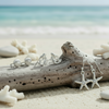 Silver-toned shell stud, mermaid stud, and starfish dangle earrings with crystal detail displayed on driftwood with white coral and beach sand and ocean in background — coastal earring set — Crabby Mermaid