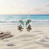 Silver-toned abalone sea turtle beach scene dangle earrings floating over beach sand with shells and driftwood and ocean in background — coastal jewelry — Crabby Mermaid