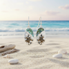 Silver-toned abalone sea turtle beach scene dangle earrings floating over beach sand with shells and driftwood and ocean in background - coastal jewelry - Crabby Mermaid