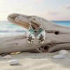Silver-toned abalone sea turtle beach scene dangle earrings leaning against driftwood with seashells on beach sand and ocean in background — coastal jewelry — Crabby Mermaid