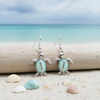 Silver-toned luminous opal sea turtle dangle earrings displayed on driftwood with pastel seashells and dreamy aqua ocean background - coastal jewelry - Crabby Mermaid