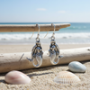 Detailed silver-toned mermaid dangle earrings hanging from driftwood with pastel shells and ocean background — coastal jewelry — Crabby Mermaid