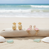 Gold-toned pineapple studs and pink enamel flamingo stud earrings displayed on driftwood with shells and sea glass on beach sand — tropical jewelry — Crabby Mermaid