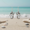 Silver-toned mermaid with starfish dangle earrings floating over beach sand with shells and sea stones and ocean waves in background — coastal jewelry — Crabby Mermaid