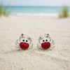 Silver-toned red glitter crab stud earrings sitting in beach sand with ocean and dunes in background — coastal jewelry — Crabby Mermaid