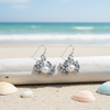 Silver-toned pearl crab dangle earrings resting on white driftwood with pastel shells on beach sand and ocean in background — coastal jewelry — Crabby Mermaid