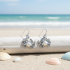 Silver-toned pearl crab dangle earrings resting on white driftwood with pastel shells on beach sand and ocean in background - coastal jewelry - Crabby Mermaid