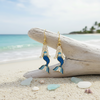 Gold-toned blue enamel mermaid dangle earrings leaning against driftwood with sea glass and shells on beach sand and tropical ocean background — coastal jewelry — Crabby Mermaid