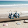 Teal patina sea turtle disc dangle earrings leaning against driftwood with pebbles and shells on beach sand and ocean in background — coastal jewelry — Crabby Mermaid