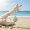 Silver-toned sea turtle teardrop necklace hanging from driftwood with sea glass and shells on beach sand and ocean in background — coastal jewelry — Crabby Mermaid