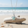 Silver-toned sea turtle pendant necklace resting on driftwood with shells on beach sand and ocean in background — coastal jewelry — Crabby Mermaid