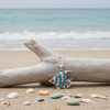 Aqua crystal angel fish pendant necklace resting on driftwood with shells and sea glass on beach sand — coastal jewelry — Crabby Mermaid