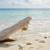 Gold-toned abalone crab necklace leaning against driftwood at water's edge with shells on beach sand — coastal jewelry — Crabby Mermaid