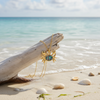 Gold-toned abalone crab necklace leaning against driftwood at water's edge with shells on beach sand - coastal jewelry - Crabby Mermaid