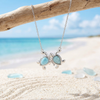 Larimar two turtle sterling silver necklace draped over driftwood on the beach with sea glass and sand - Crabby Mermaid