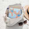 Starfish coastal cosmetic bag tucked inside white canvas tote bag on Florida beach sand with straw hat and sunglasses - Crabby Mermaid