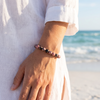 Woman wearing black stripe rhodonite gemstone beaded bracelet on the beach in a white linen shirt with ocean waves in the background — Crabby Mermaid