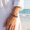 Woman wearing black stripe rhodonite gemstone beaded bracelet on the beach in a white linen shirt with ocean waves in the background — Crabby Mermaid