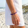 Woman wearing amethyst gemstone beaded bracelet on the beach in a white linen dress with sea oats and ocean waves in the background — Crabby Mermaid