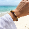 Woman wearing polychrome jasper gemstone beaded bracelet at the beach in a white linen shirt with teal ocean waves in the background — Crabby Mermaid