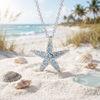 Sterling silver starfish necklace with aqua Swarovski crystals on white sand with seashells at Florida beach — Crabby Mermaid