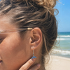 Sterling silver whale tail earrings with sapphire crystals worn by sandy blonde model on Florida beach — handcrafted in Ireland — Crabby Mermaid