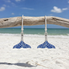Sterling silver whale tail drop earrings with sapphire blue crystals hanging from driftwood on Florida beach — handcrafted in Ireland — Crabby Mermaid