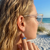 Close-up of sterling silver whale tail earrings with sapphire blue crystals on sandy blonde model at Florida Gulf Coast beach — Crabby Mermaid