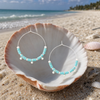 Turquoise stone disc hoop earrings in silver displayed in a white seashell on a Florida Gulf Coast beach — Crabby Mermaid