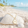 Gold epoxy whale tail charm necklace resting on white shell on Florida Gulf Coast beach with sea oats and turquoise water