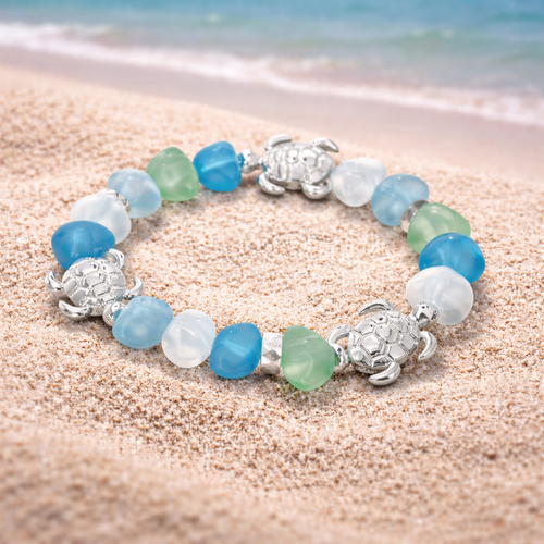 Sea glass turtle stretch bracelet displayed on sand with ocean in background - coastal jewelry - Crabby Mermaid