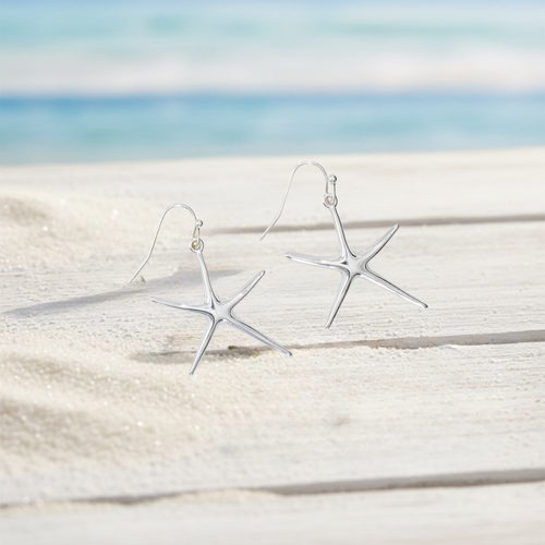 Polished silver starfish dangle earrings with French wire hooks displayed on weathered white wood with sand and ocean background - coastal jewelry - Crabby Mermaid