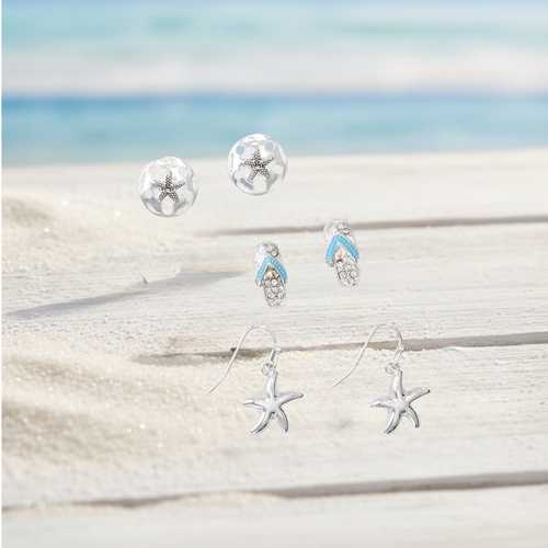 Silver sand dollar studs, crystal flip flop studs, and starfish dangle earrings displayed on weathered white wood with sand and ocean background — coastal earring set — Crabby Mermaid